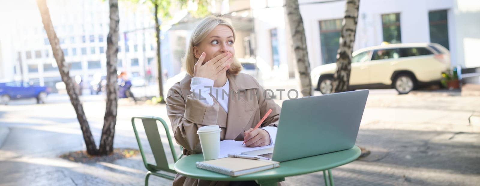 Image of woman sitting with laptop outside on street, using laptop, looking surprised, amazed by something she saw online.