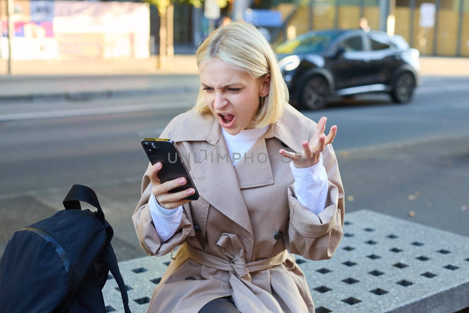 Portrait of angry young woman, shouting at her mobile phone in frustrated, being outraged, sitting on street bench outdoors.