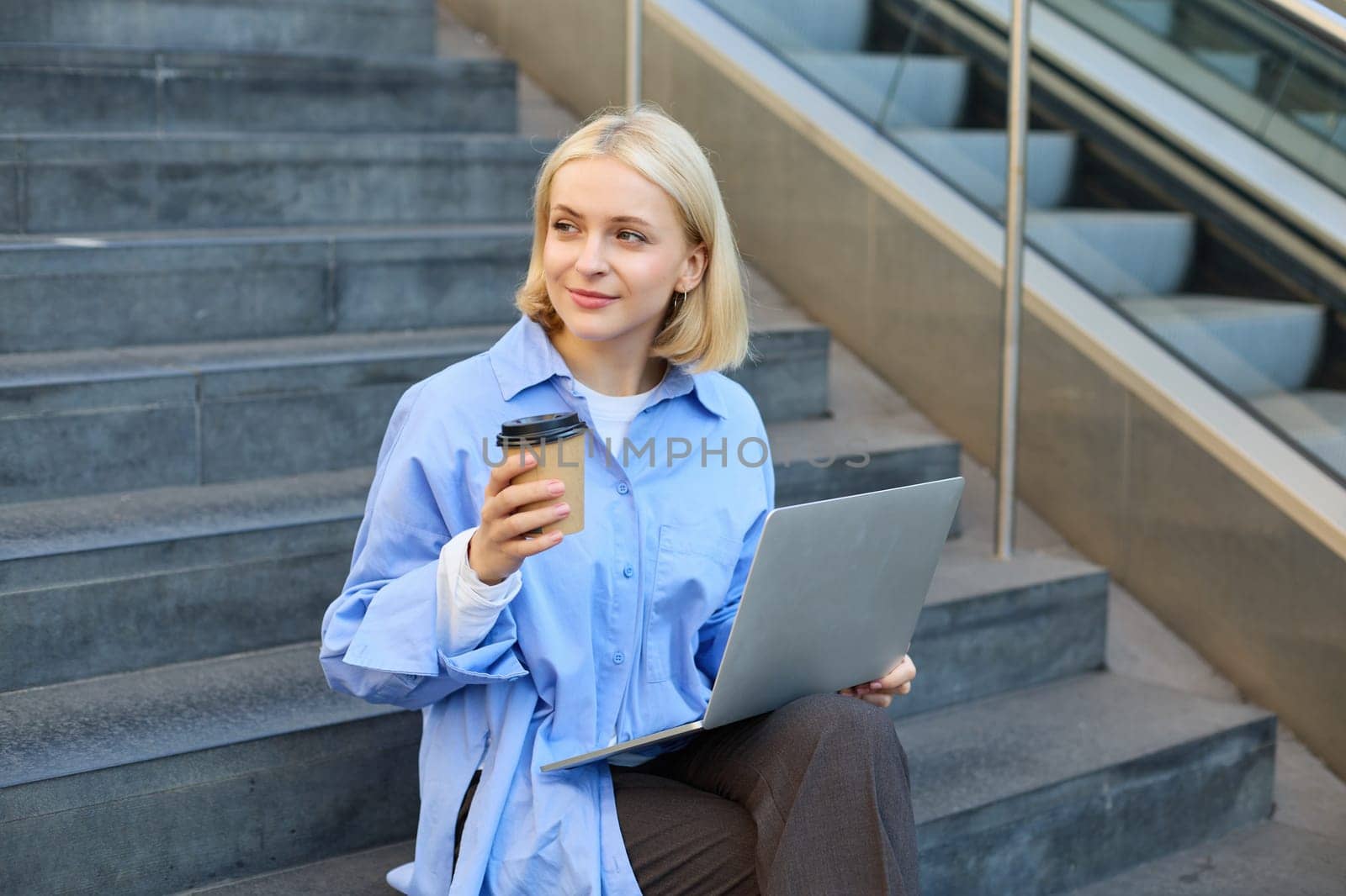 Smiling young urban woman, student working on laptop, sitting outdoors on stairs, drinking coffee, studying, e-learning on fresh air.