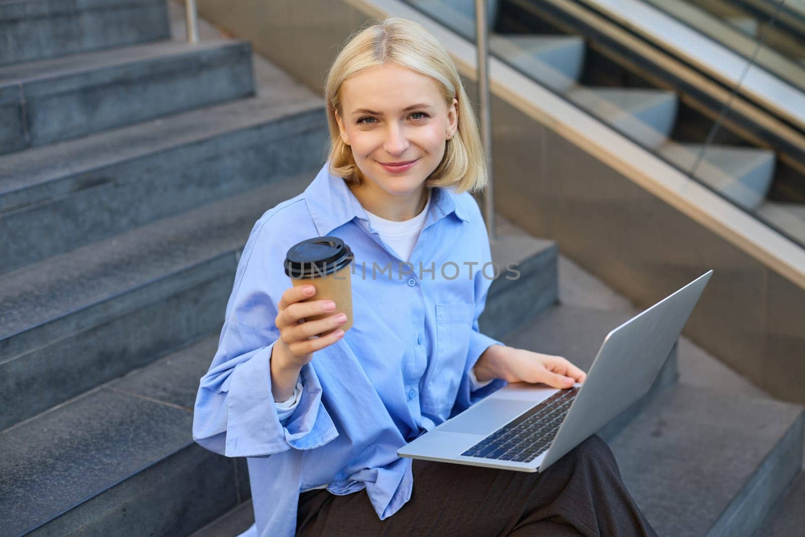 Image of stylish young woman, college student with laptop, sitting on city stairs and drinking coffee, working on project, studying outdoors, connects to public wifi, enjoying weather.