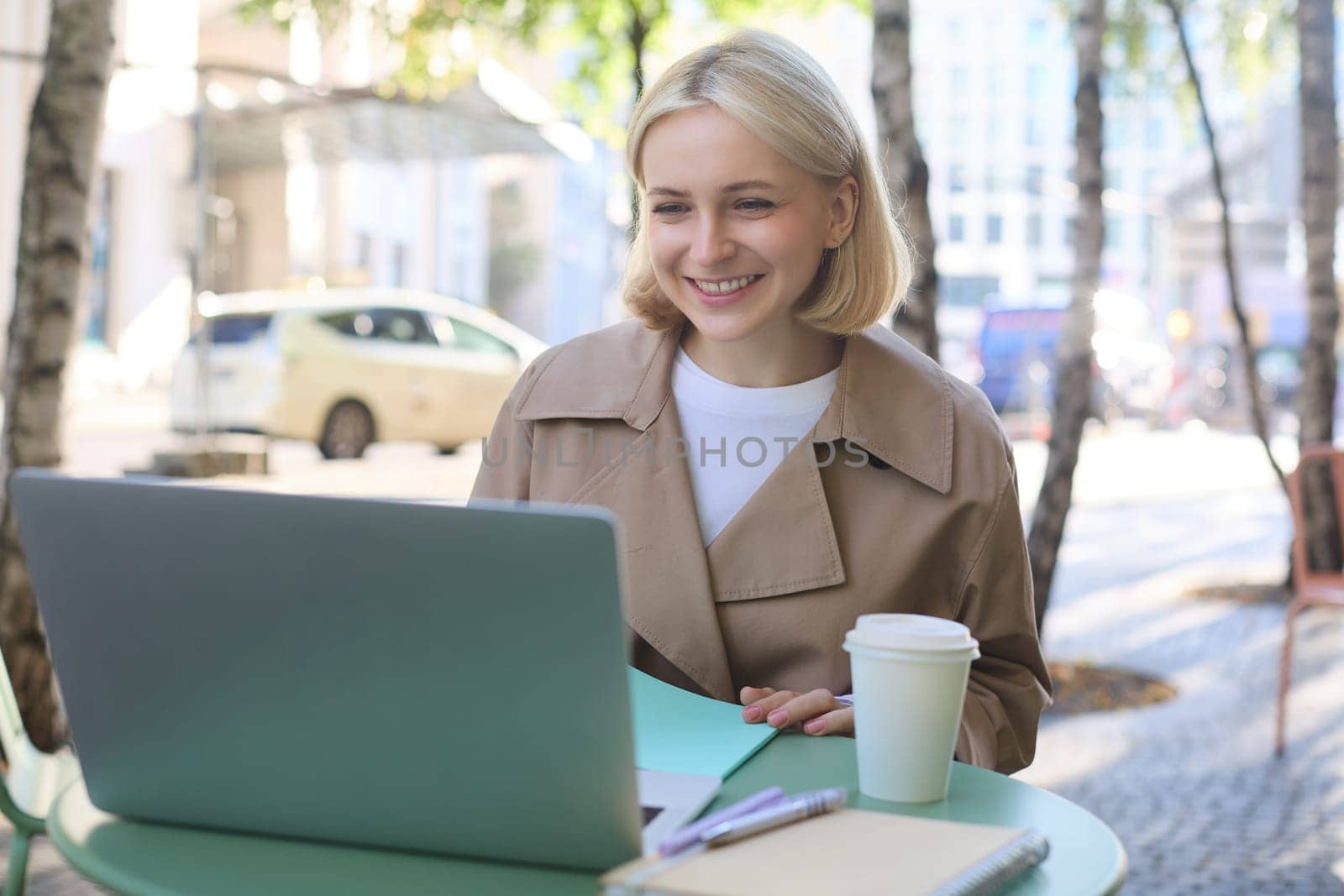 Young woman, freelancer, student doing homework in outdoor cafe, drinking her coffee on street, using laptop, connects to online lecture or doing course in internet.