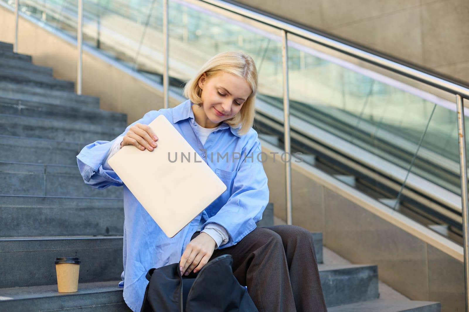 Image of stylish young woman, putting away her laptop in backpack, sitting on stairs near campus, packing for university, drinking coffee.