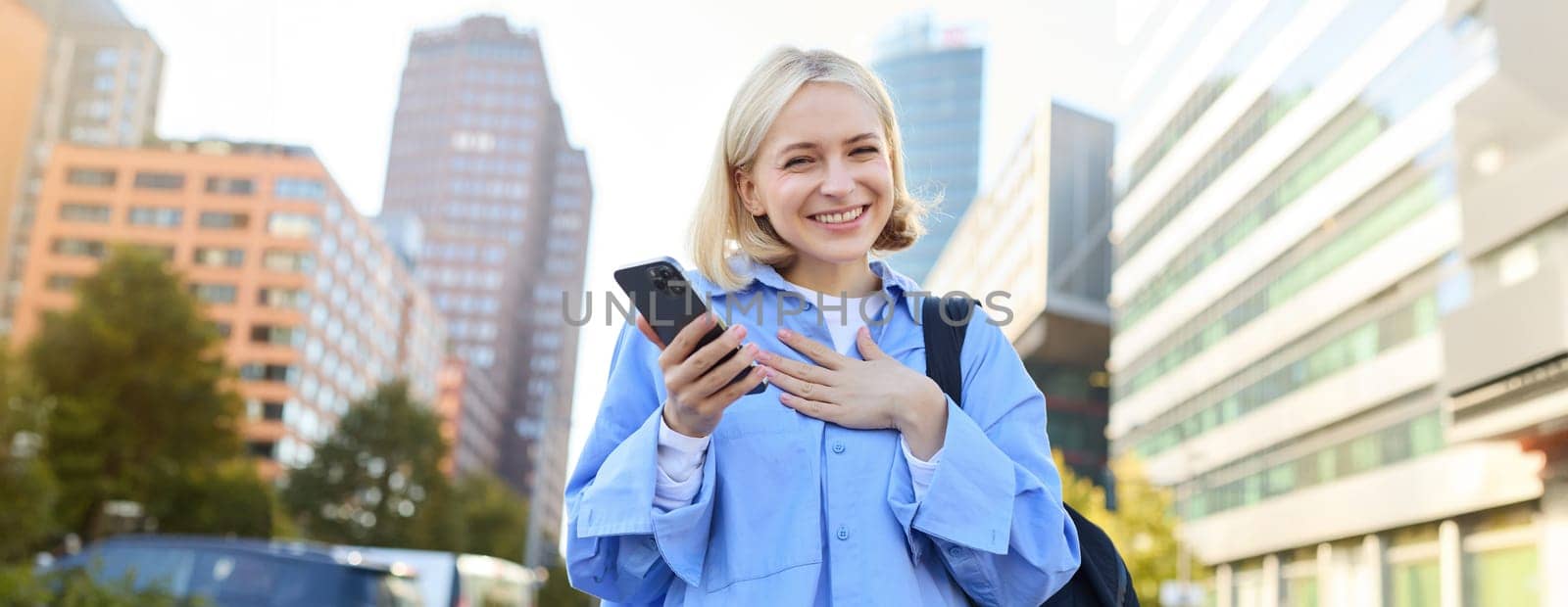 Close up portrait of stylish young blonde woman, standing on street, checking her mobile phone, using smartphone app to get around town, looking at online map for directions by Benzoix