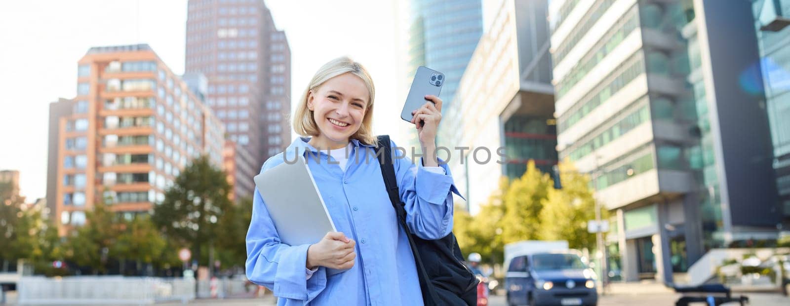 Image of young stylish, modern blond woman in blue shirt, holding laptop and backpack, using smartphone, smiling, walking along the street, posing in city centre.