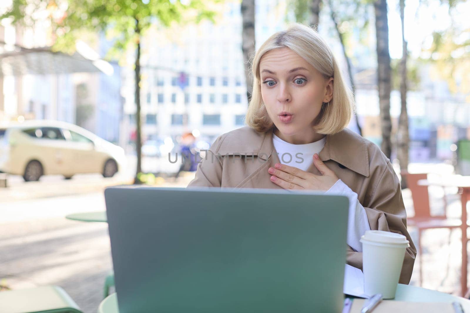 Close up portrait of young blond woman, sitting outdoors with laptop, drinking coffee in cafe, looking amazed and surprised at device screen, hears awesome news by Benzoix