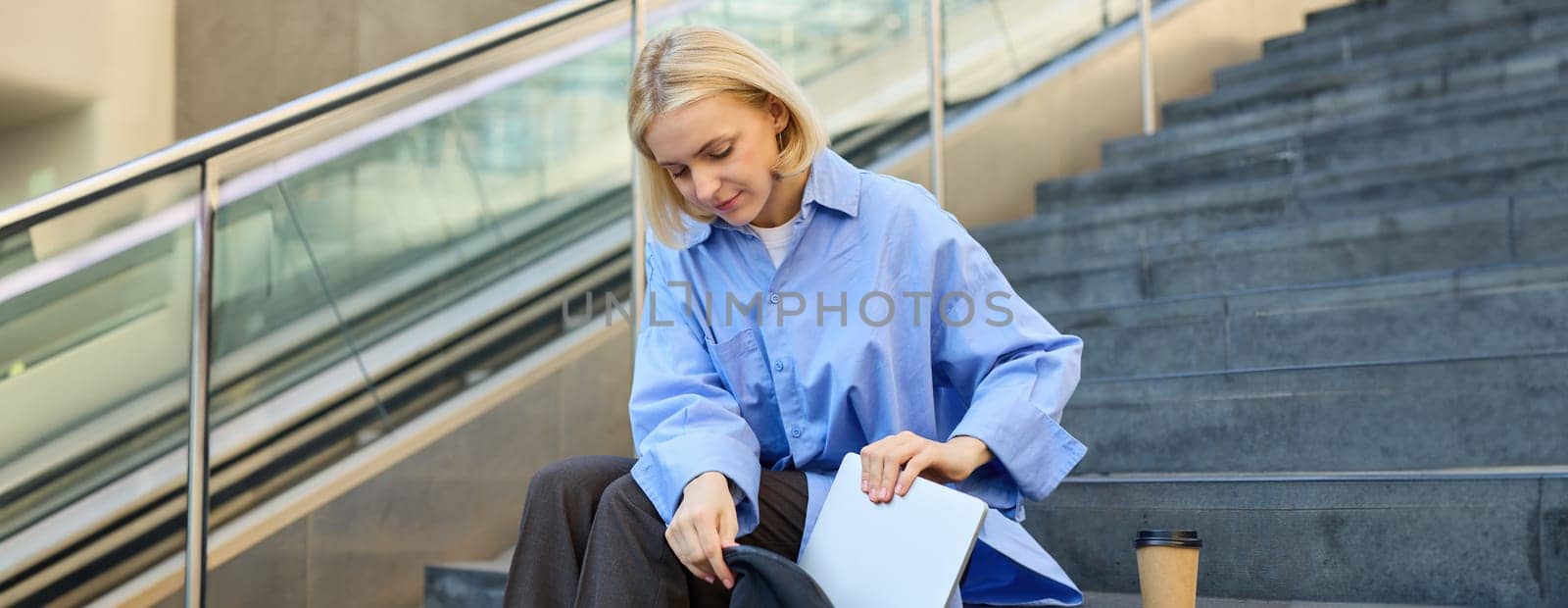 Young modern woman sits on stairs outdoors, drinks coffee, packs her laptop inside backpack. Lifestyle and student concept