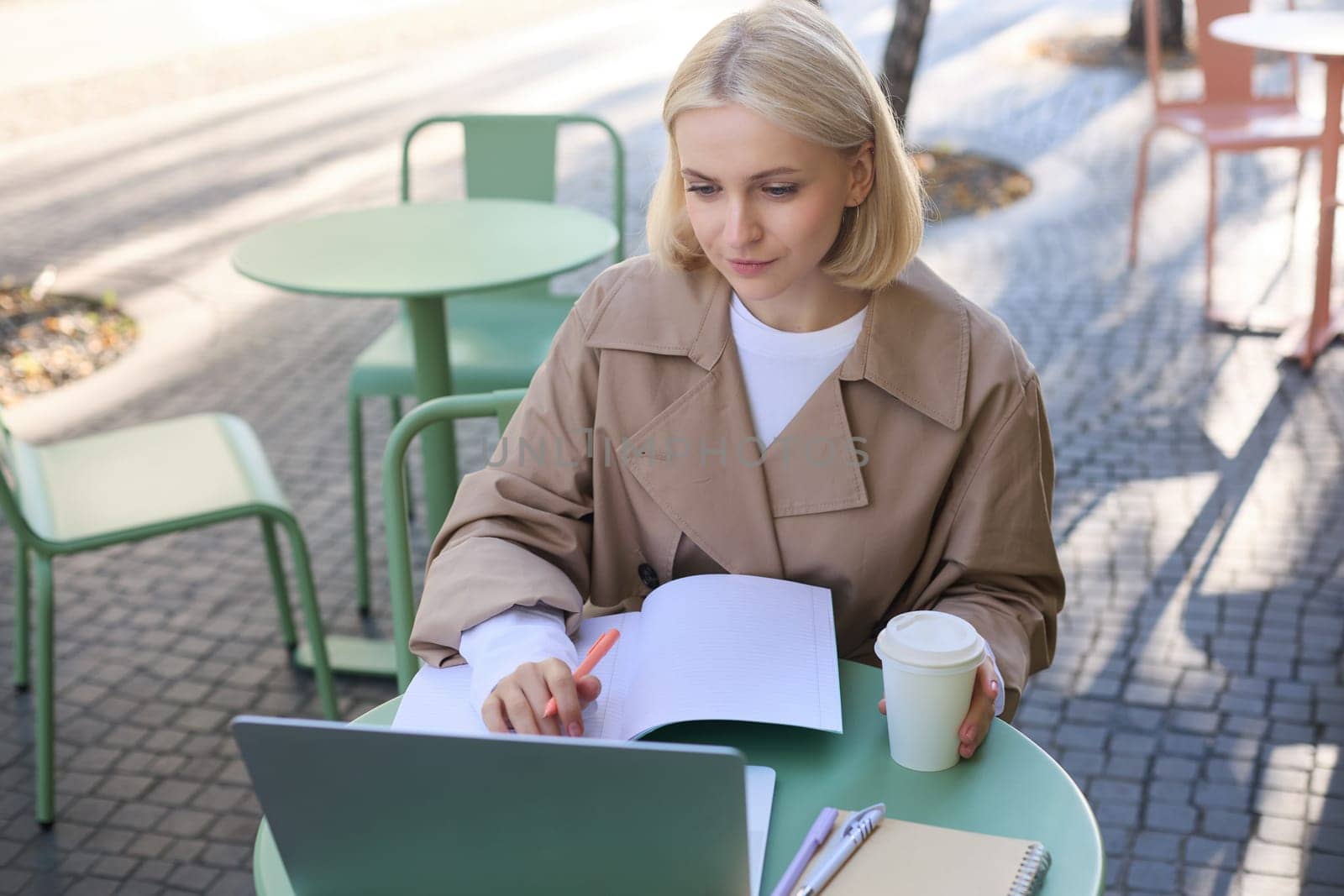 Image of young woman studying outdoors, sitting in cafe, making notes while listening to online lecture using laptop, making notes in notebook, writing.
