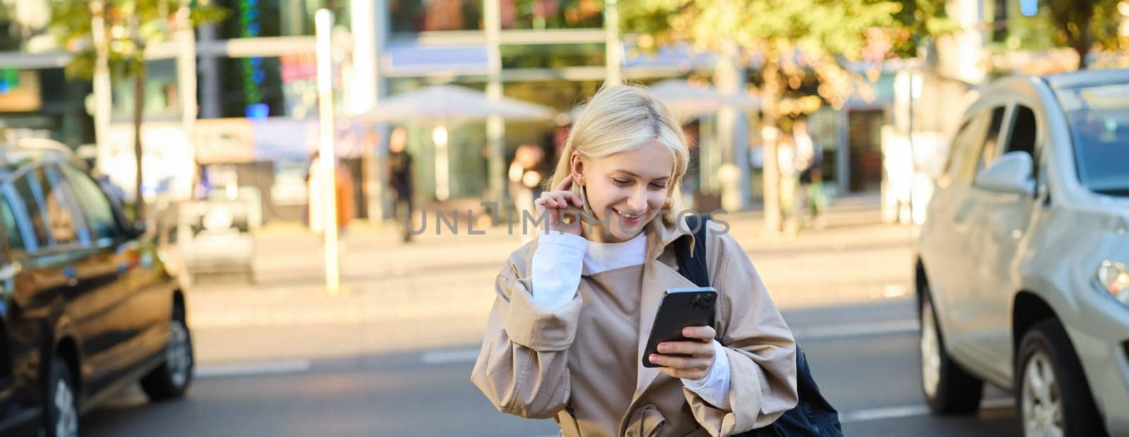 Modern young woman on street, reading message on mobile phone, using smartphone, standing near busy road and smiling.