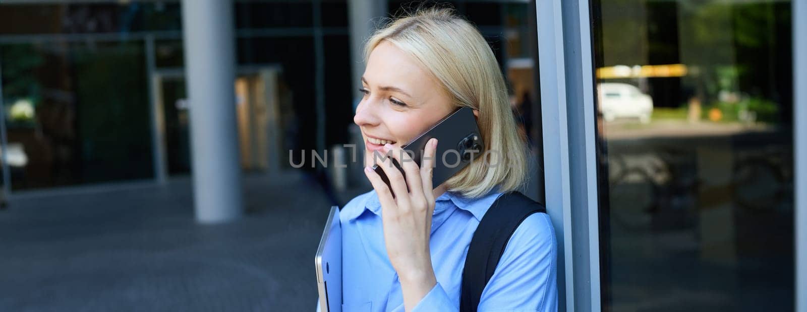 Close up portrait of modern young woman, student standing near building in city centre, talking on mobile phone, having chat on smartphone and smiling by Benzoix