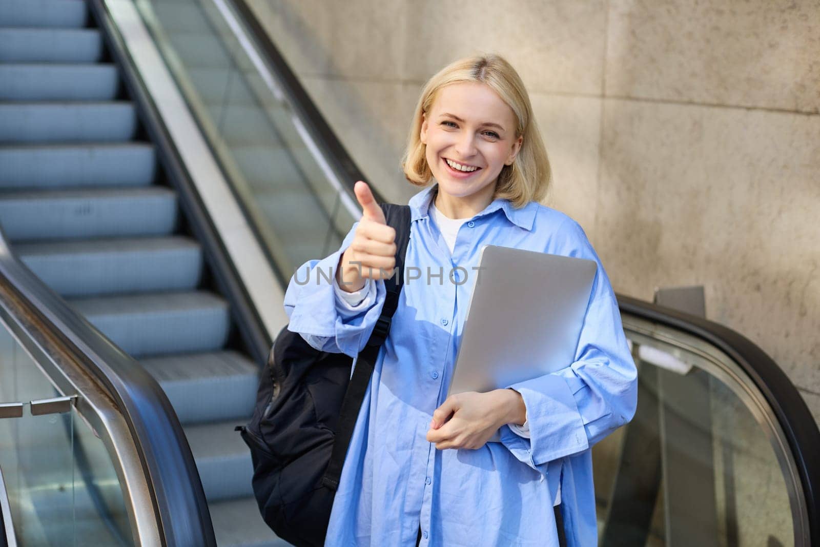 Friendly, smiling young woman, college student with laptop and backpack, using escalator, showing thumbs up in approval, recommending product or company by Benzoix