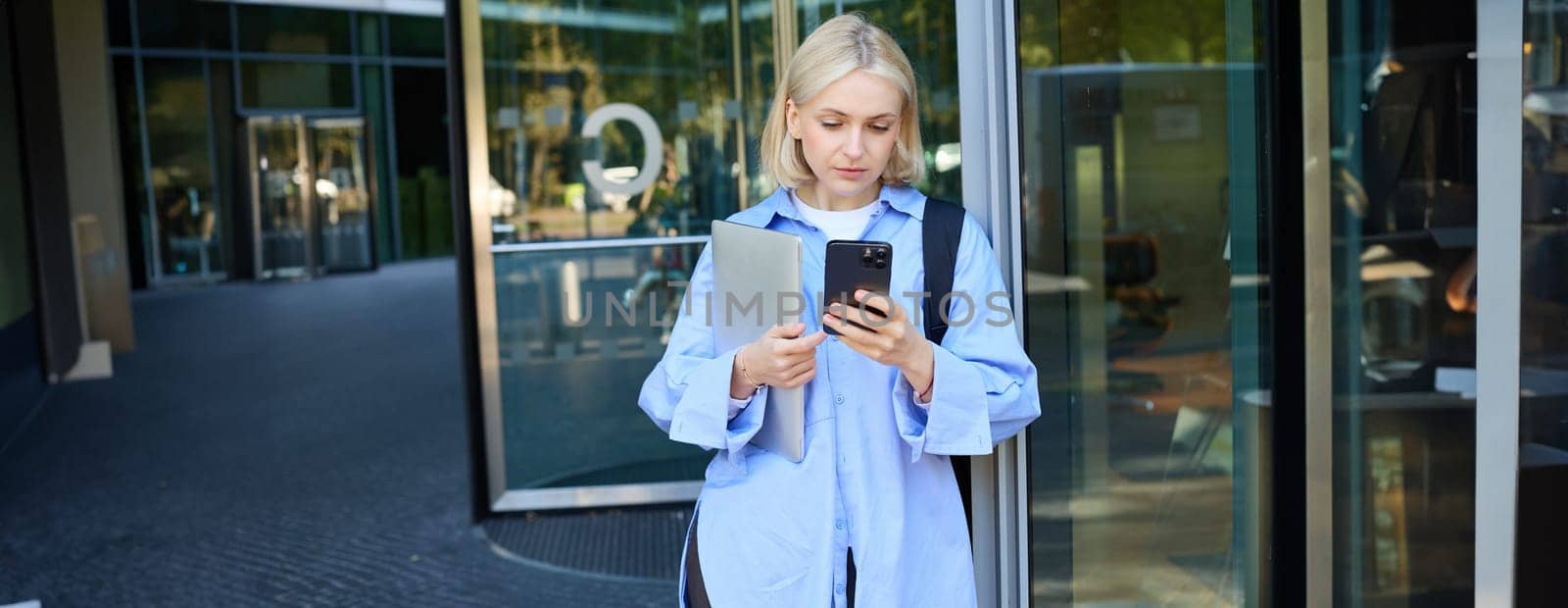 Image of young blond woman in blue shirt, holding laptop, waiting for someone near office building, using smartphone, mobile phone application.