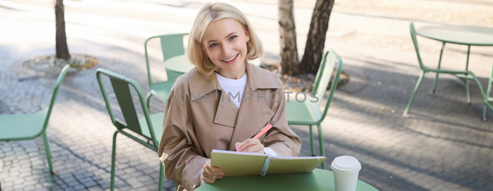 Image of young smiling woman, sitting in outdoor cafe, doing sketches, drawing art on street, spending time on coffee shop and creating art.