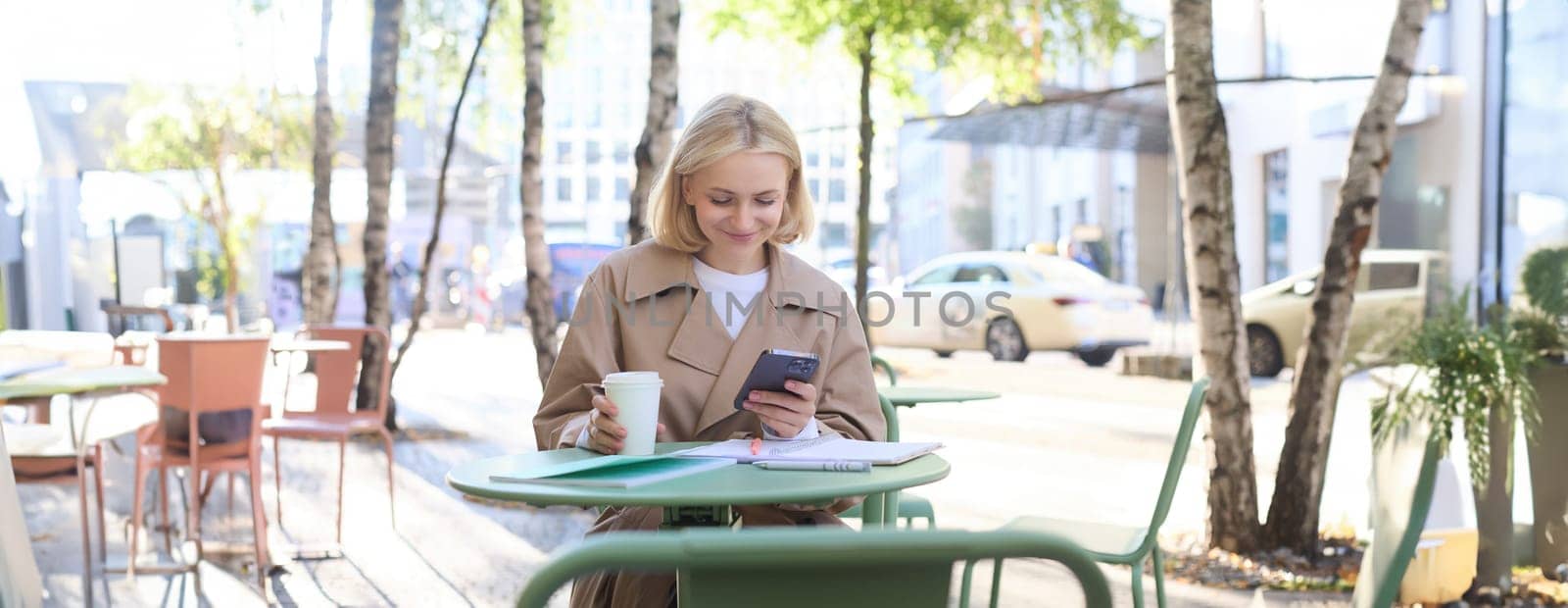 Image of young woman drinking coffee, sitting in outdoor cafe with smartphone, sending message, reading notification on mobile phone and smiling by Benzoix