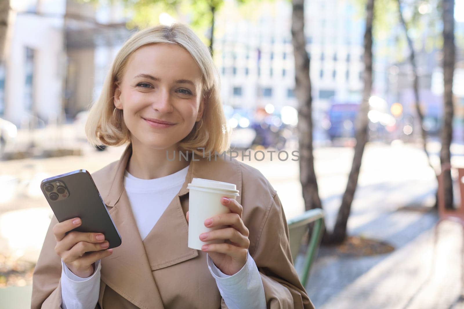 Portrait of cute blond woman with smartphone, drinking chai, coffee at favourite place in city centre, sitting outdoors, holding mobile phone by Benzoix