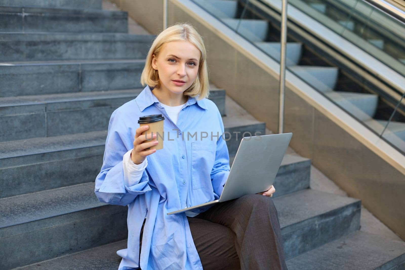 Portrait of young urban woman sitting on stairs with cup of takeaway coffee and working on laptop, studying outdoors on fresh air, freelancing.