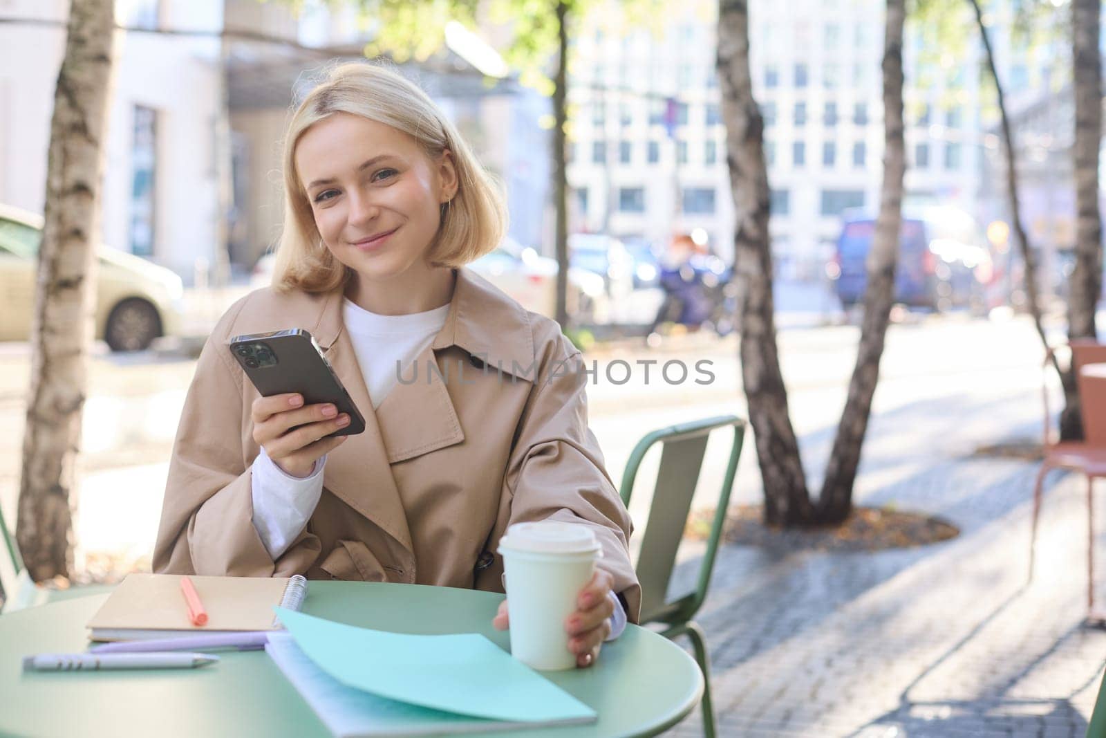 Portrait of blond smiling woman with smartphone, holding cup of coffee, drinking chai and enjoying sunny day outdoors in city centre.