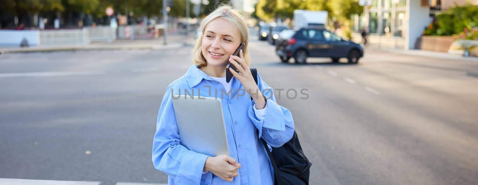 Close up portrait of young woman with laptop, standing on street near busy road in city, talking on mobile phone, answers a call by Benzoix
