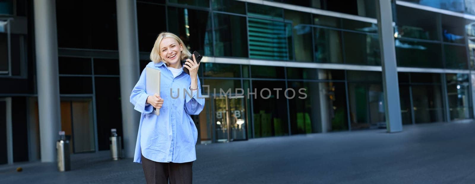 Vertical shot of modern, stylish young blond woman smiling, posing in trousers and blue collar shirt, holding smartphone and laughing, posing near campus building, has laptop in hand.