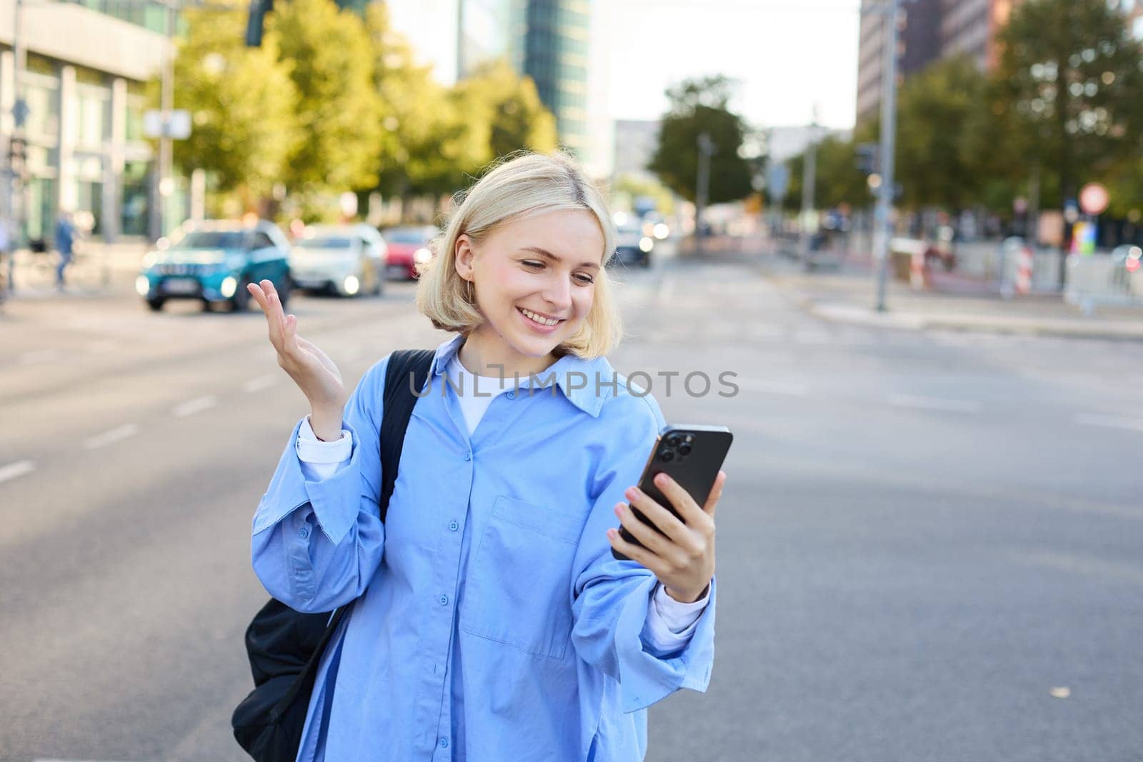 Portrait of young office worker, woman walking along street, chatting on the phone, looking at smartphone and showing smth behind her, video chats, connects to online conversation via mobile app.
