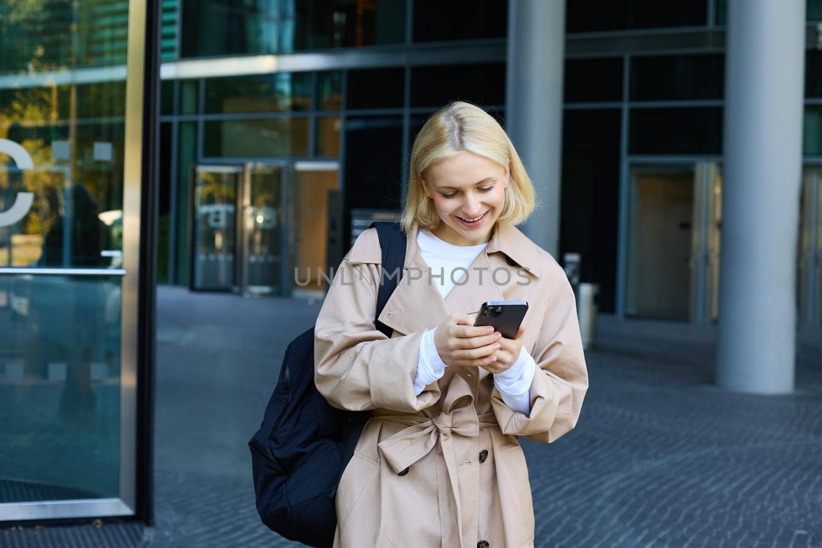 Street photo of young blond woman with smartphone, walking in city, waiting for someone, order taxi on mobile application.