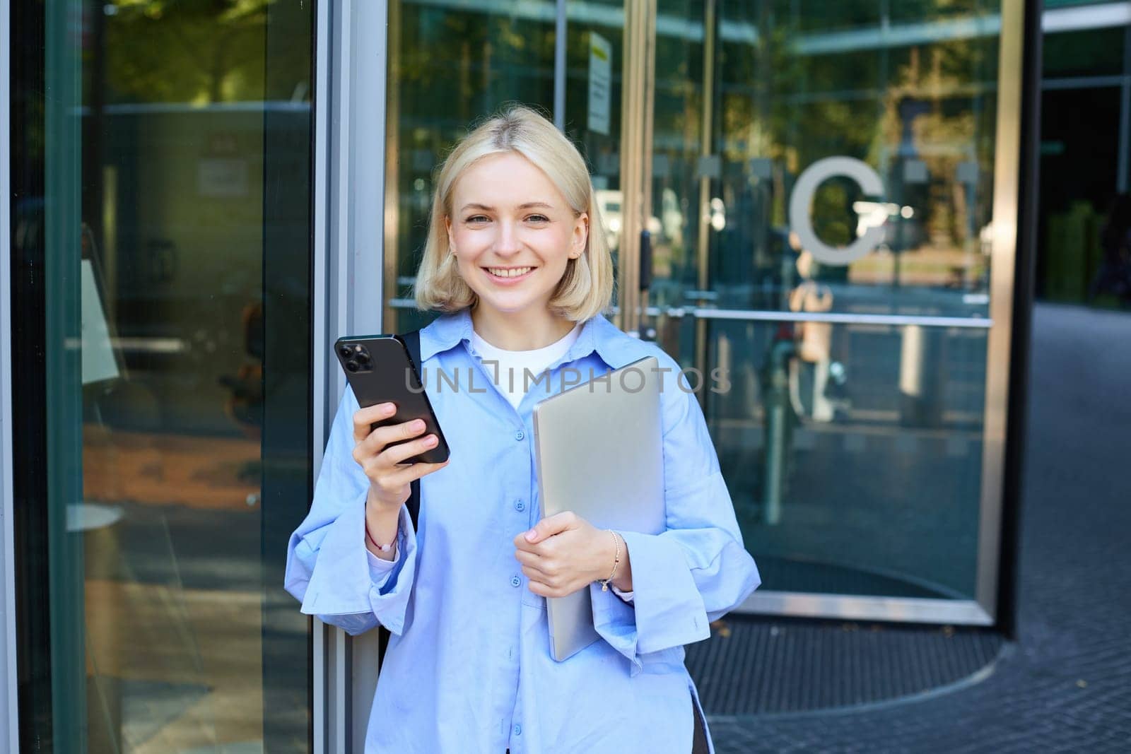 Image of stylish young woman, student walking out of the college campus, holding smartphone and laptop, has backpack on shoulder, waiting for someone outside with mobile phone by Benzoix