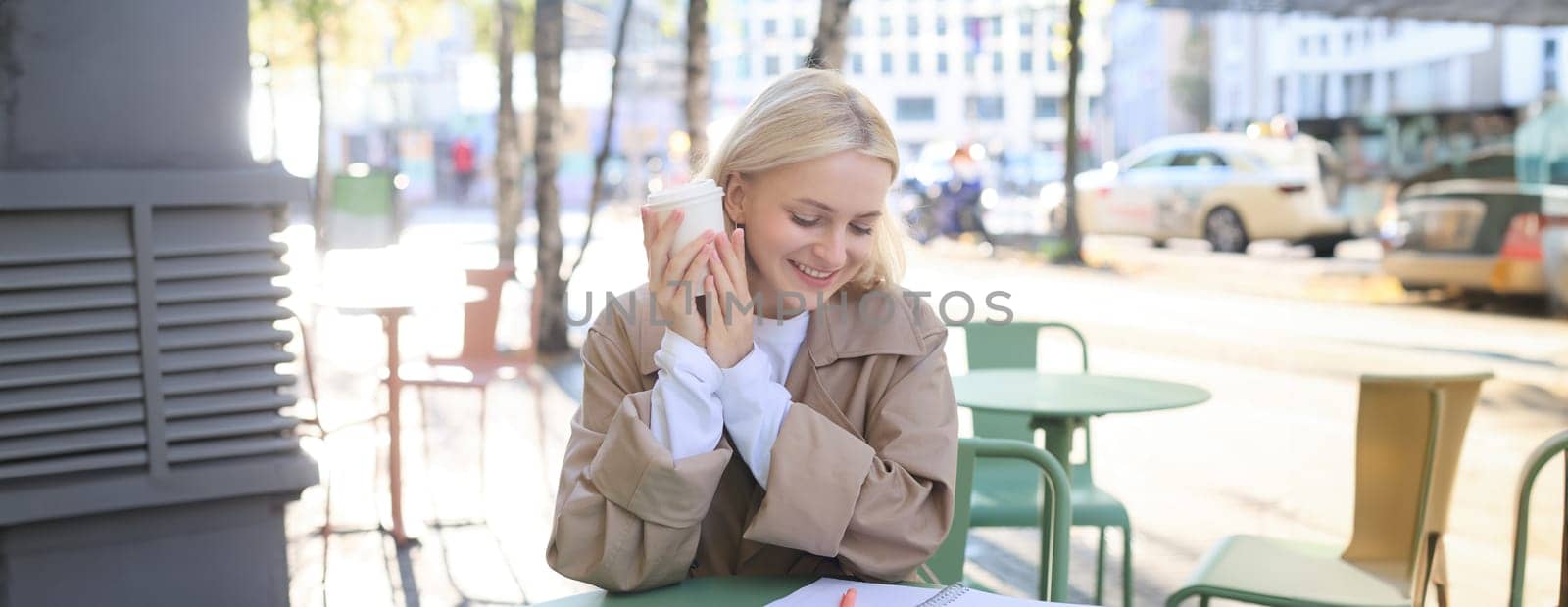 Beautiful blond girl doing homework in street cafe, holding takeaway cup, drinking coffee, spending time outdoors in city centre.