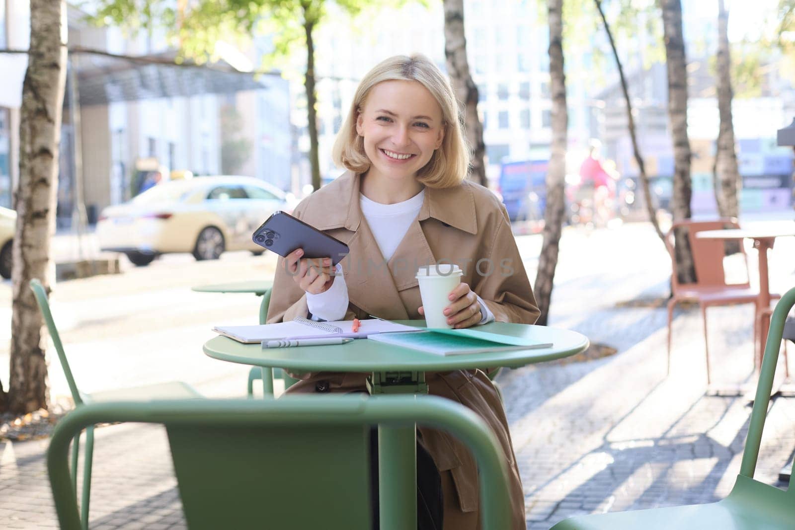 Portrait of modern young woman in city centre, sitting in cafe outdoors, drinking coffee, using mobile phone, smiling by Benzoix