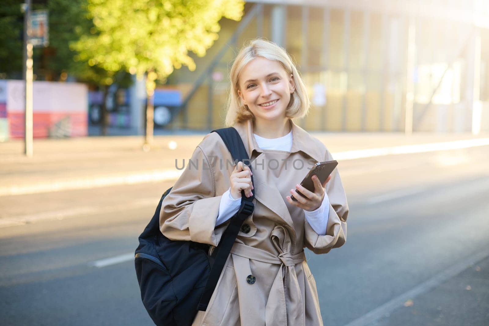 Street style portrait of blonde smiling woman walks on street, sunny bright day, holding smartphone, carries backpack, orders taxi on mobile application, sends message on cell phone by Benzoix
