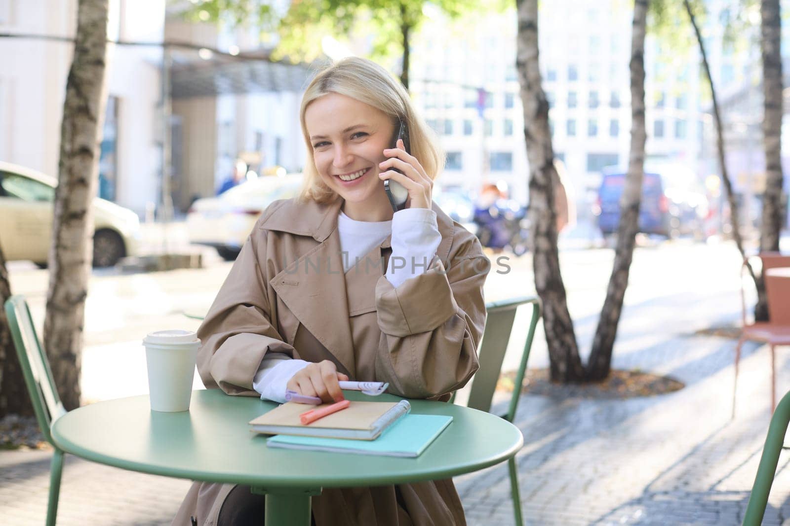 Lifestyle and urban people concept. Young smiling girl, student sitting in cafe outdoors, talking on mobile phone, chatting with friend or coworker over the telephone, studying or doing homework by Benzoix
