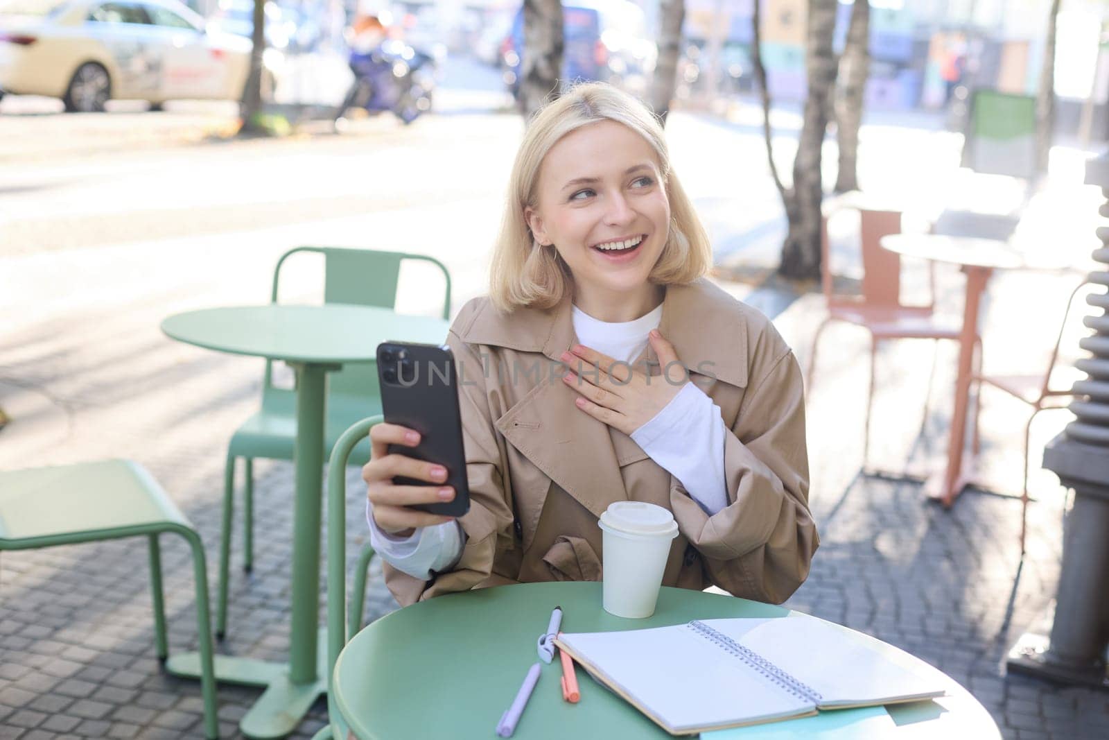 Portrait of young stylish woman in trench, sitting outdoors in city cafe, talking on video chat, using mobile phone to connect to online conversation, drinking coffee.