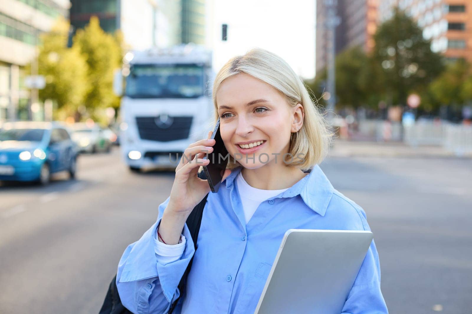 Close up portrait of young woman with laptop, standing on street near busy road in city, talking on mobile phone, answers a call. Lifestyle concept