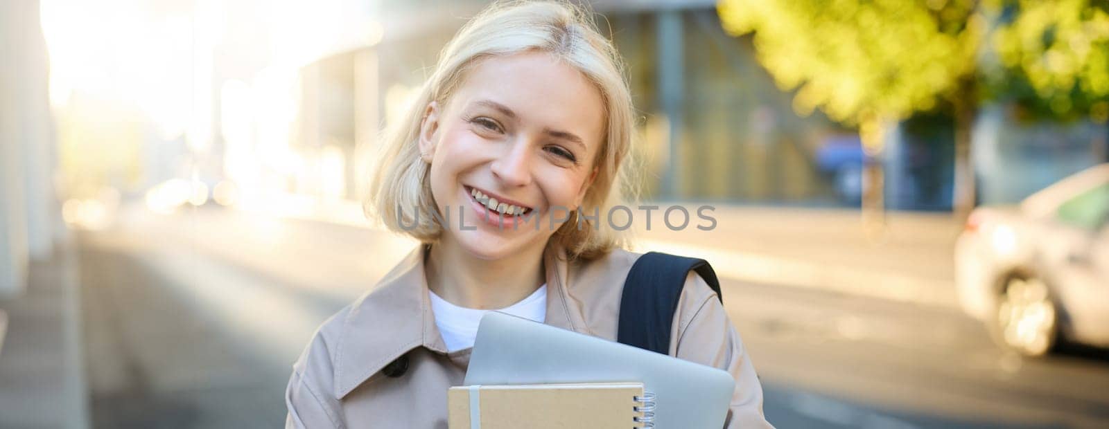 Close up portrait of smiling blond woman, standing on street with notebooks, carries journal and work documents, looking happy at camera by Benzoix