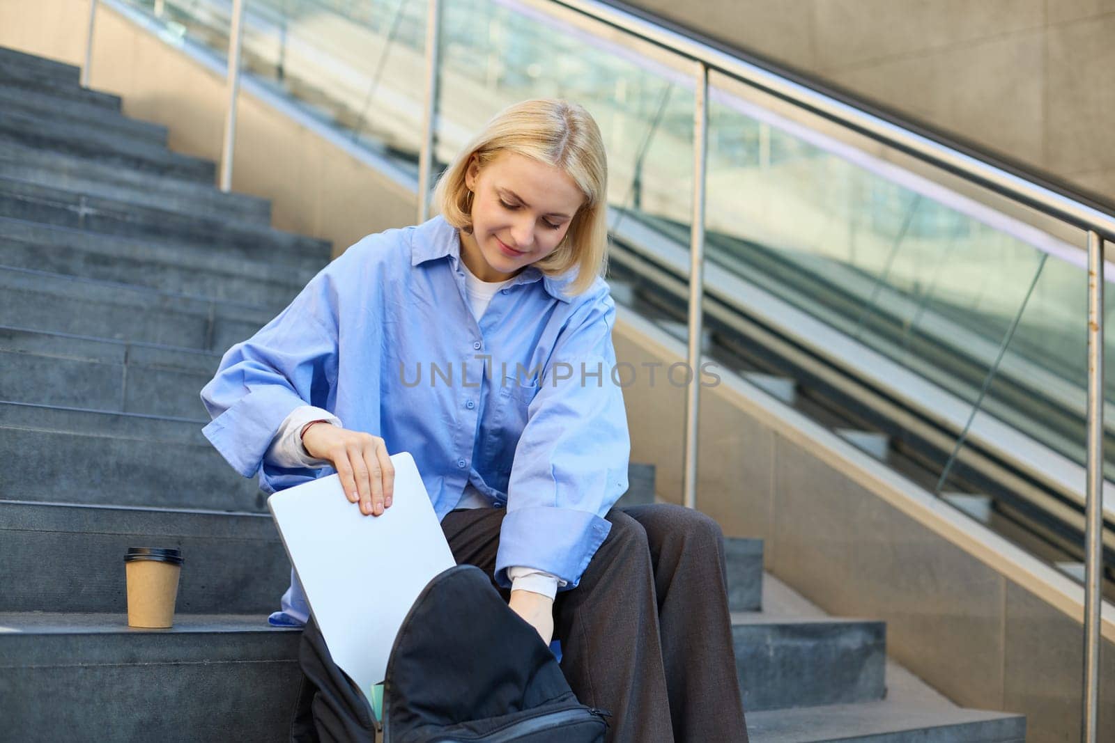 Portrait of young modern woman, student freelancer, sitting on public stairs outdoors, putting away her laptop, packing backpack, drinking coffee outside.