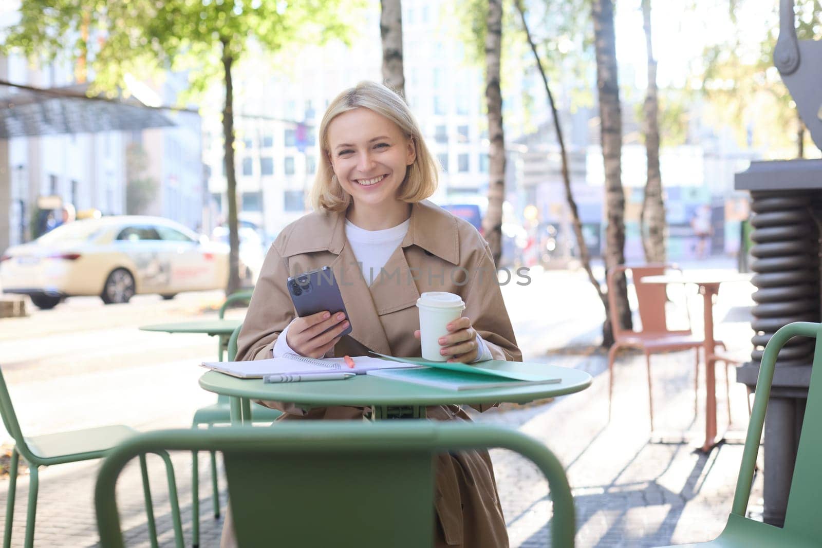 Portrait of young woman studying, sitting in outdoor cafe with smartphone and journals, doing homework, wearing trench coat on sunny day.