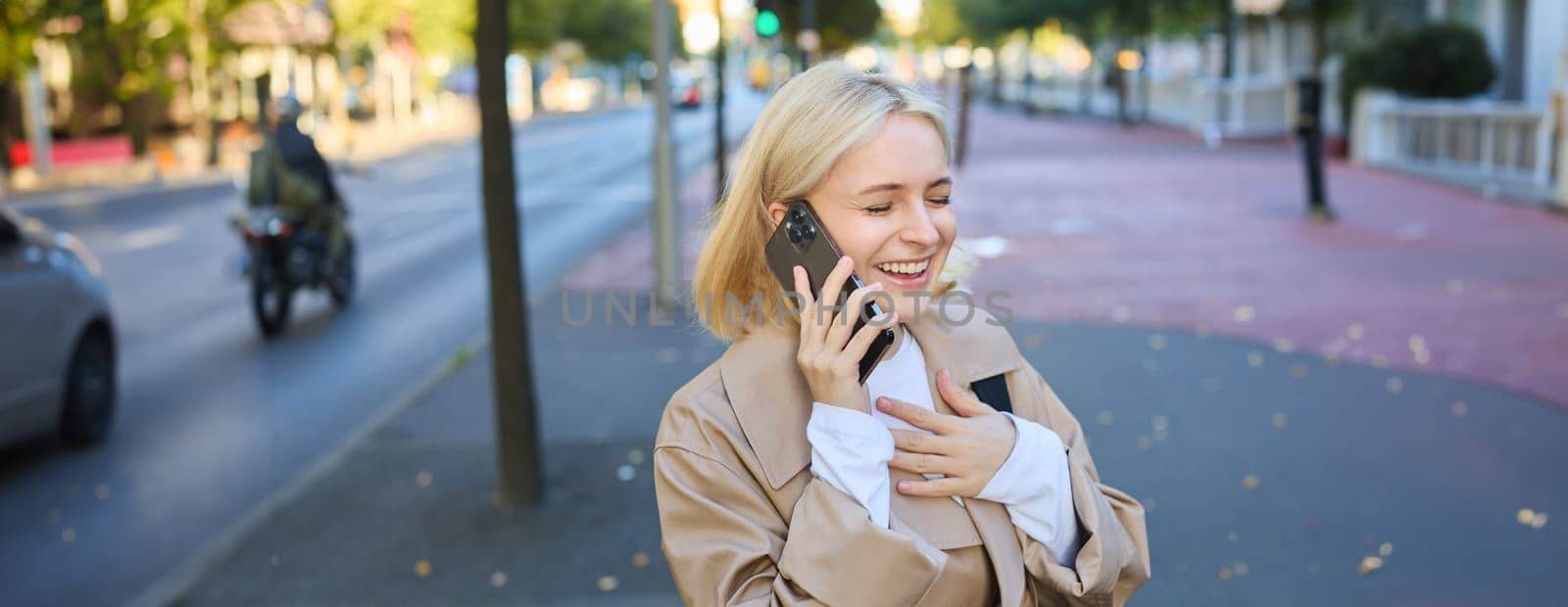 Close up portrait of beautiful young woman, blonge girl walking on street with mobile phone, chatting with friend, has happy face expression while talking over cellphone on her way.