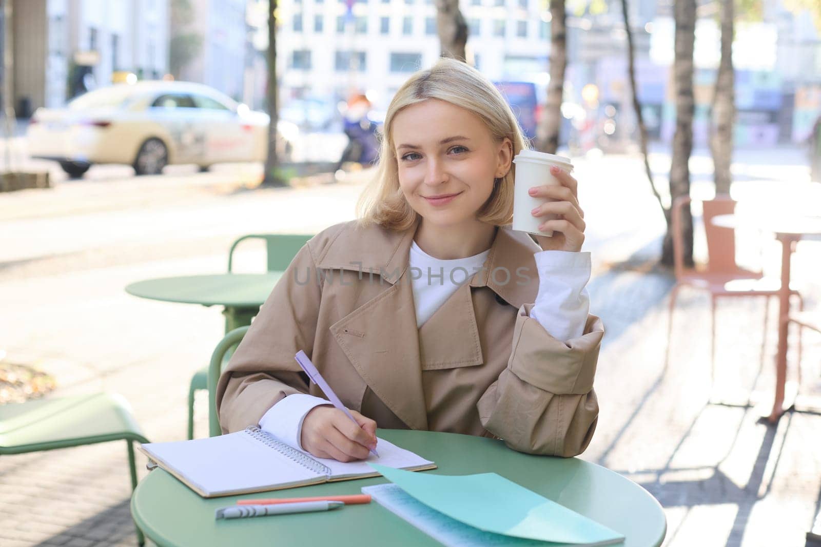 Image of young smiling woman, female entrepreneur writing, making corrections in documents, using notebook, drinking coffee and sitting in outdoor cafe.