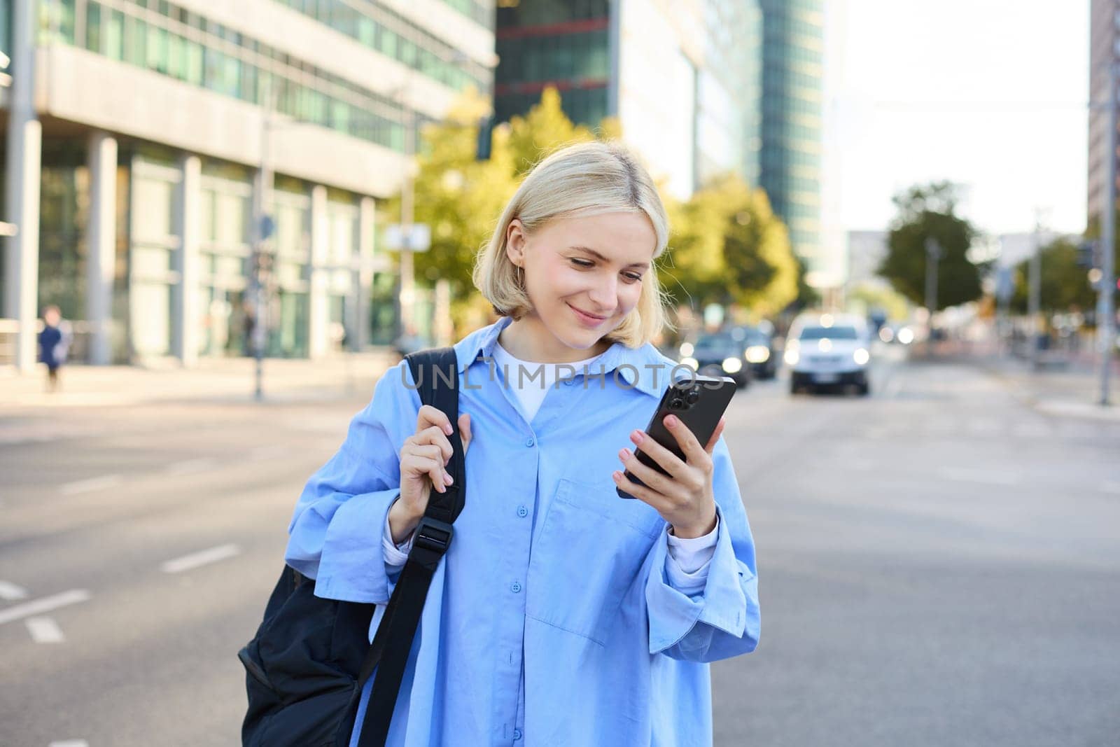 Portrait of young city girl, urban woman with backpack, reading messages on smartphone and smiling, checking notifications, standing on street.