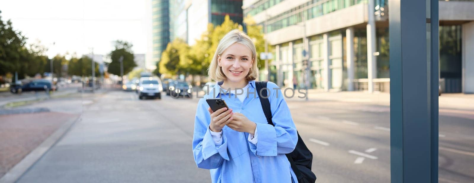 Street style shot of young smiling woman with smartphone, standing on street, bright sunny day, road with cars behind her back, using mobile phone, waiting for someone outdoors.