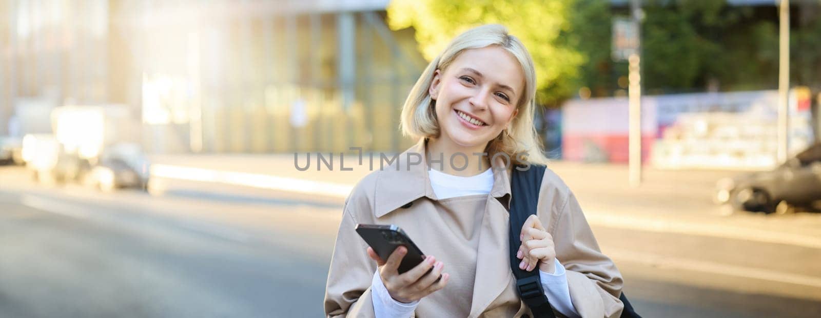 Close up portrait of young smiling college girl, student with backpack, holding smartphone, looking happy at camera, waiting on street.