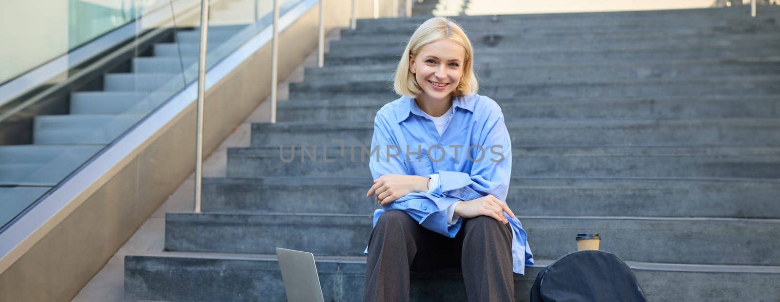 Urban style portrait of young woman, student sitting on city street stairs, with backpack, cup of coffee and laptop, looking happy and upbeat, smiling.