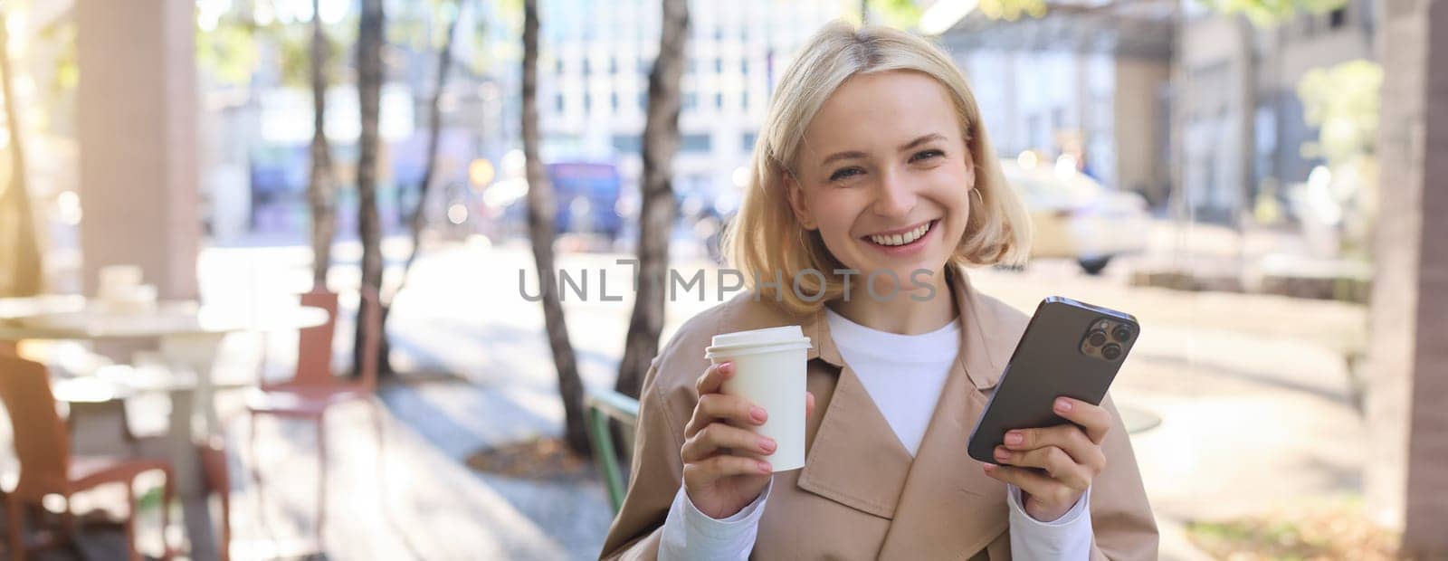 Portrait of blond smiling woman with smartphone, holding cup of coffee, drinking chai and enjoying sunny day outdoors in city centre by Benzoix