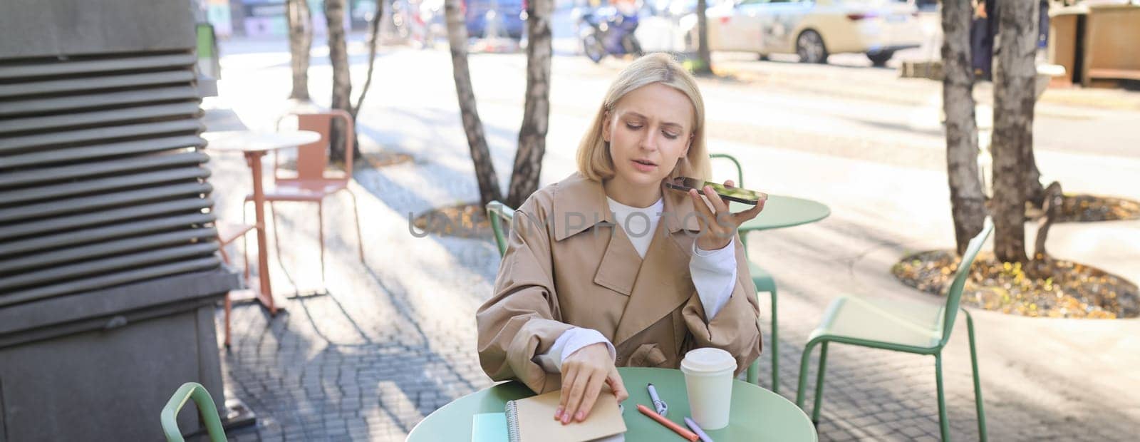 Portrait of serious, concentrated woman, opens her notebook, records voice message, sending notification to coworker, drinking coffee, working outdoors.