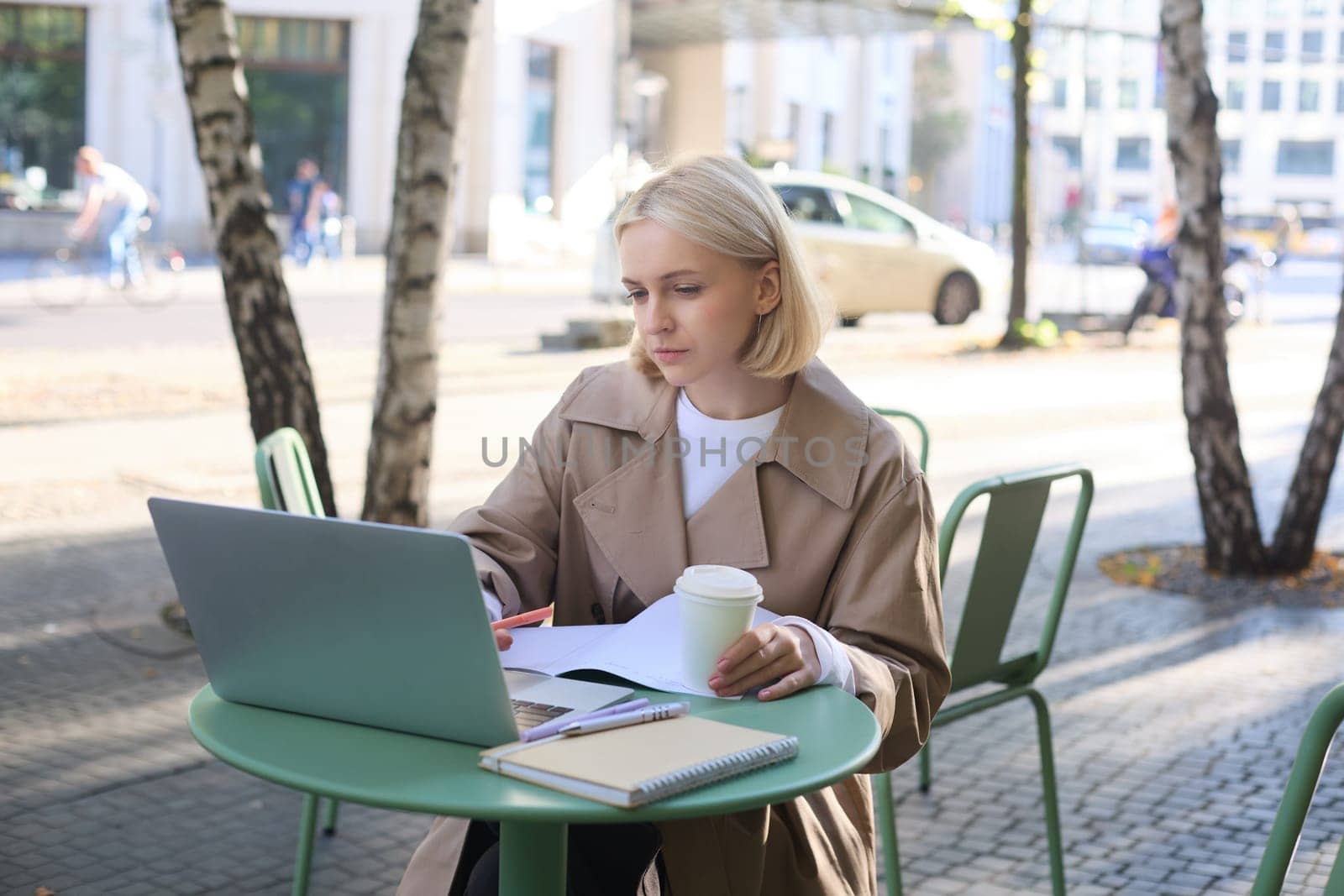 Image of young modern woman, sitting in cafe outdoors, drinking coffee or tea, looking at laptop, working on project, freelancing, making notes in notebook. Lifestyle concept