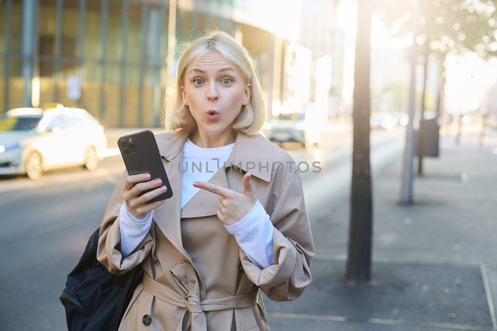 Image of surprised girl on street, pointing at mobile phone and gasping amazed, saying wow. Technology and urban lifestyle concept