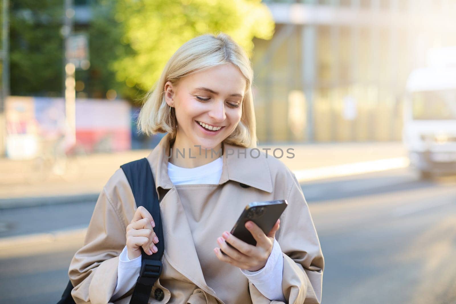 Close up portrait of happy, beautiful young woman with backpack, standing on street and using mobile phone, waiting for taxi near road, smiling by Benzoix