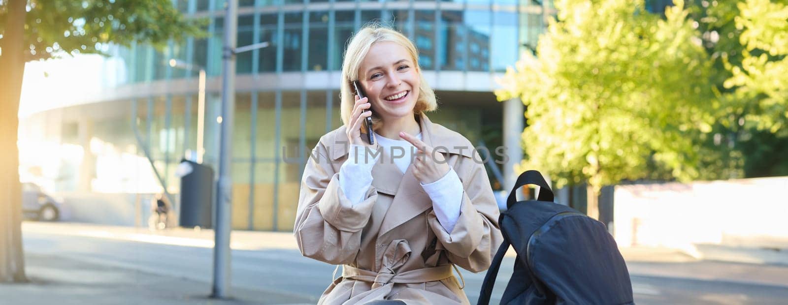 Portrait of cheerful smiling woman, talking on mobile phone, pointing at her smartphone while calling someone, smiling and laughing at camera, sitting on street bench outdoors by Benzoix