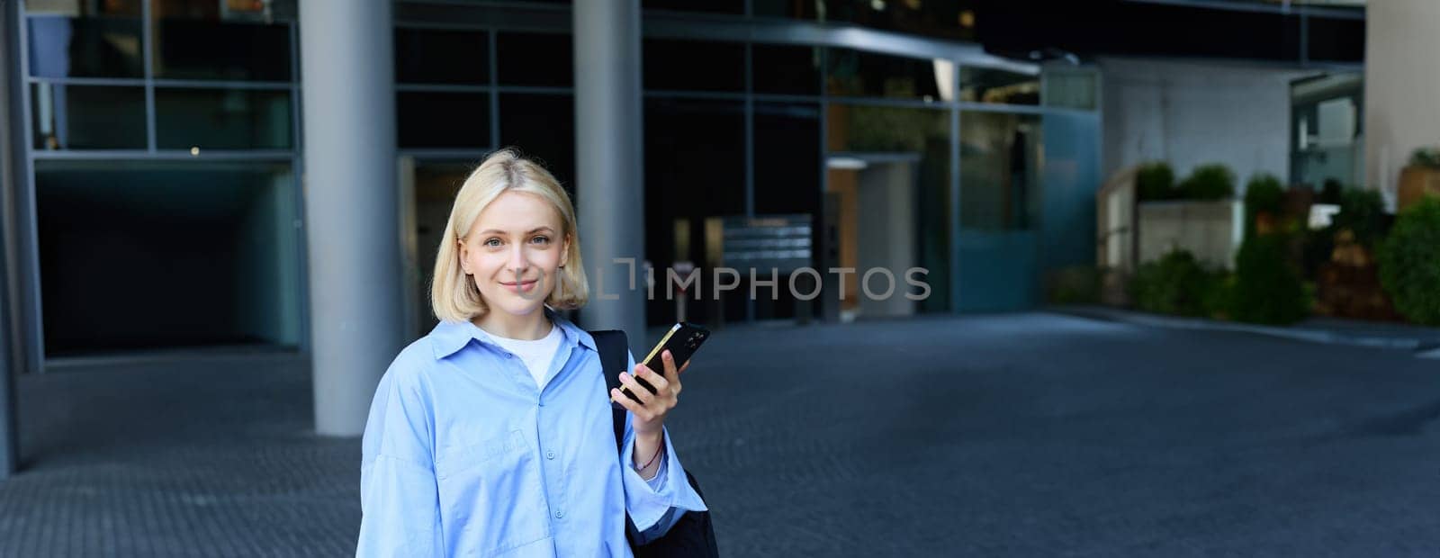 Vertical portrait of young office manager, woman in blue collar shirt and backpack, holding laptop and smartphone, waiting near business building on street, smiling at camera by Benzoix