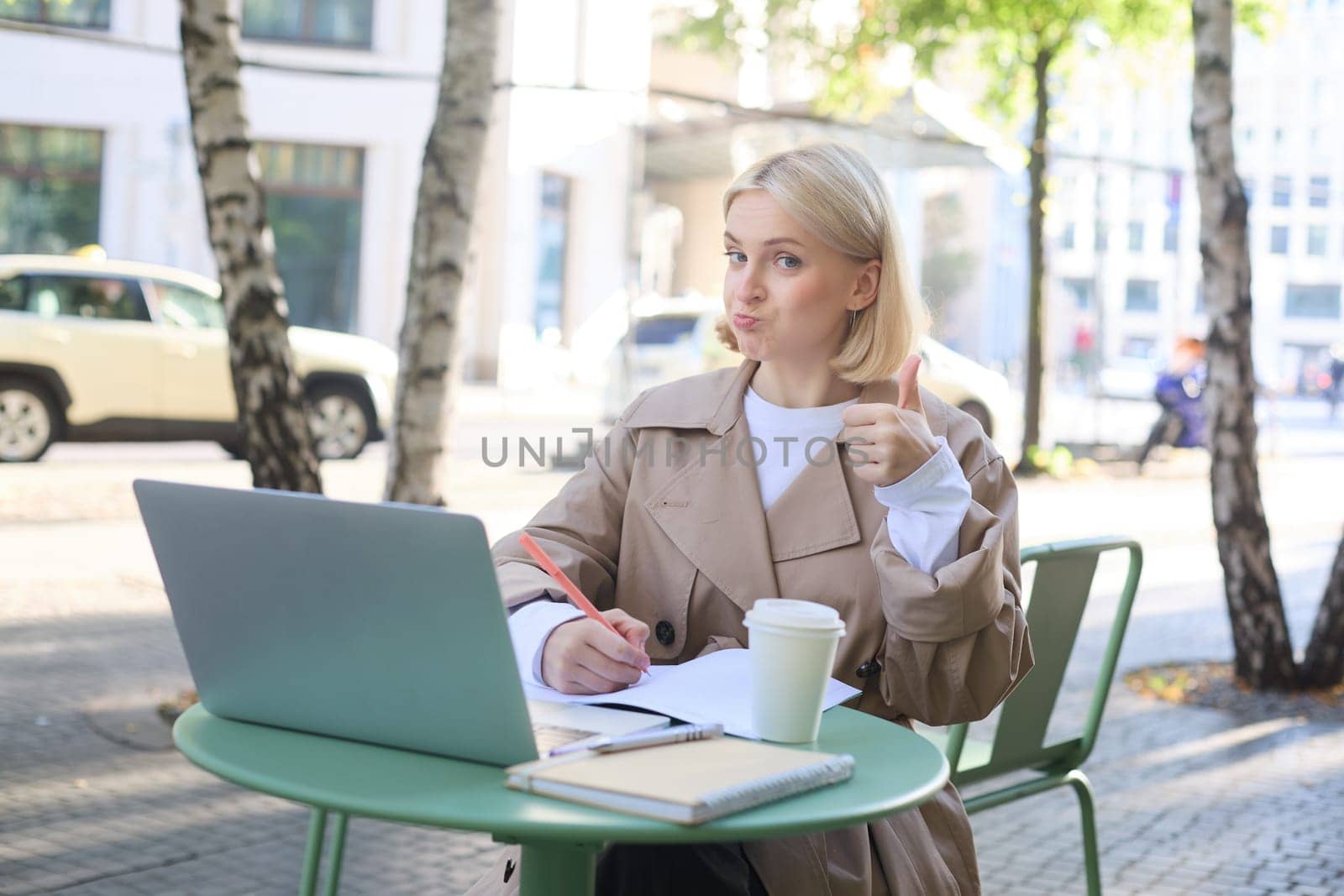 Image of impressed, satisfied young woman, sitting in outdoor cafe with laptop, showing thumbs up and nod in approval. Lifestyle concept
