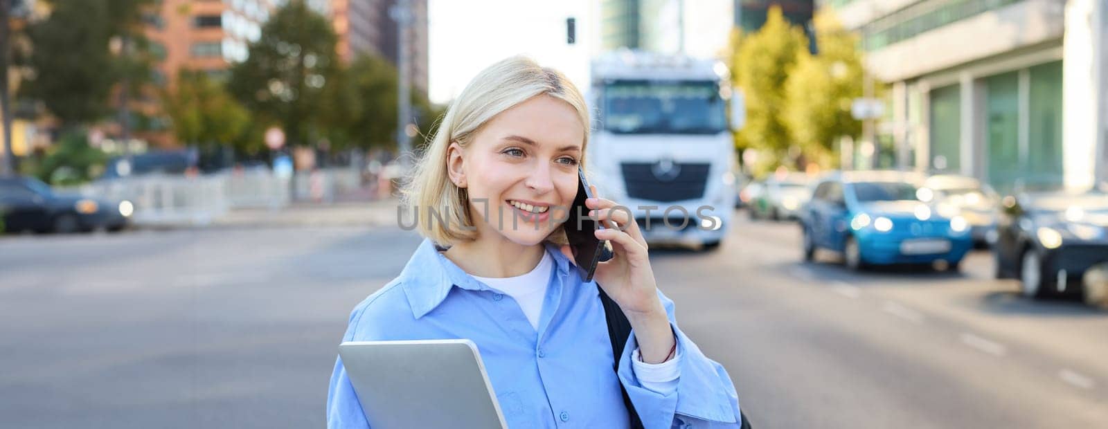 Close up portrait of young woman with laptop, standing on street near busy road in city, talking on mobile phone, answers a call. Lifestyle concept