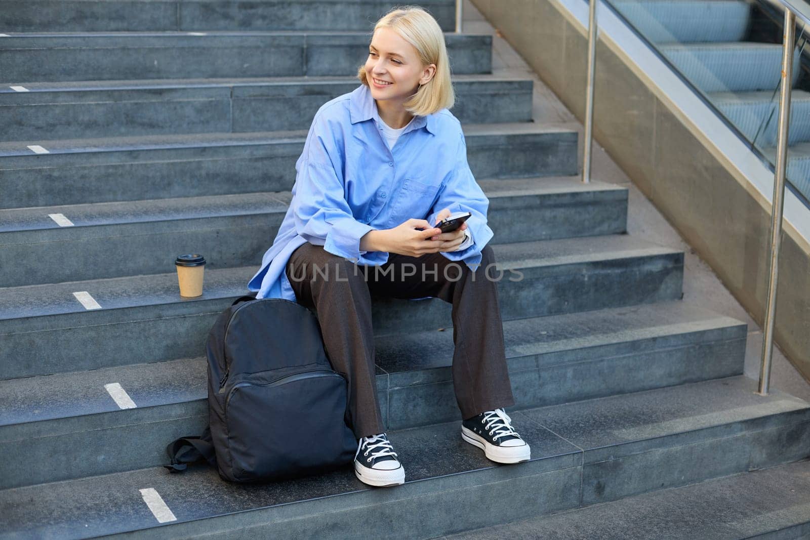 Portrait of woman on stairs, sitting with backpack, using mobile phone, messaging, scrolling social media on lunch break in university by Benzoix